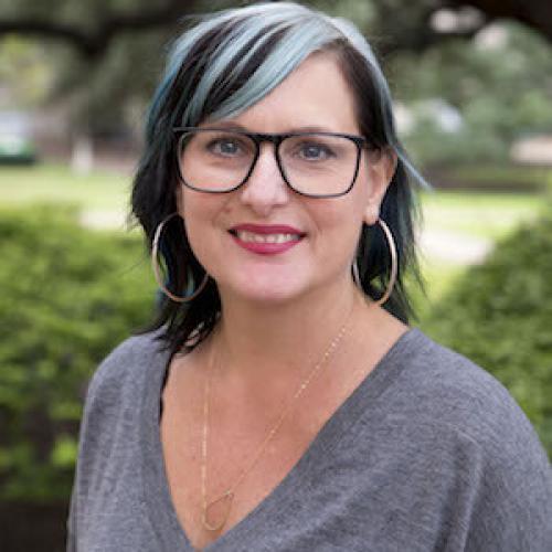 White women wearing a grey shirt, hoop earrings, and black glasses