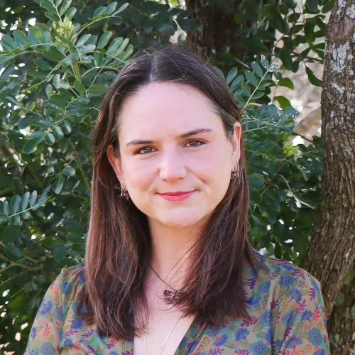 Michelle has long brown hair and wears a multi-colored blouse in front of green foliage.